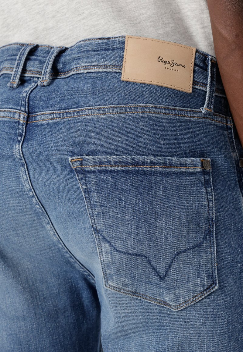 Vista trasera de una persona que lleva vaqueros de mezclilla azules con una etiqueta de cuero de Pepe Jeans London encima del bolsillo trasero derecho.