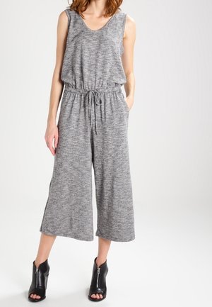 Nainen pukeutuneena hihattomaan harmaaseen neulottuun haalariin, jossa on kiristysnauha vyötäröllä ja leveät lyhytlahkeiset housut, yhdistettyinä mustiin avokkaisiin nilkkureihin.