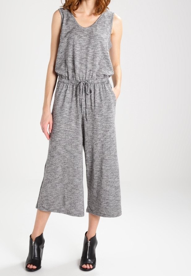 Femme portant une combinaison sans manches en maille grise avec taille à cordon de serrage et jambes larges raccourcies, associée à des bottines noires à bout ouvert.