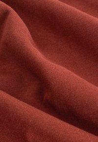 Nahaufnahme eines strukturierten, rostfarbenen Stoffes mit sanften Falten und Rillen, die seine glatte, gewebte Oberfläche und den warmen Farbton hervorheben.