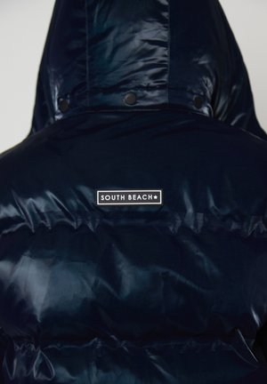 Giacca a vento lucida di colore blu scuro con cappuccio, caratterizzata da un patch "South Beach" sulla parte superiore della schiena.