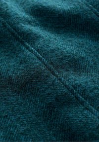 Tessuto di lana verde scuro con una consistenza morbida e pelosa, caratterizzato da un delicato motivo a scacchi, con una cucitura visibile che corre diagonalmente.