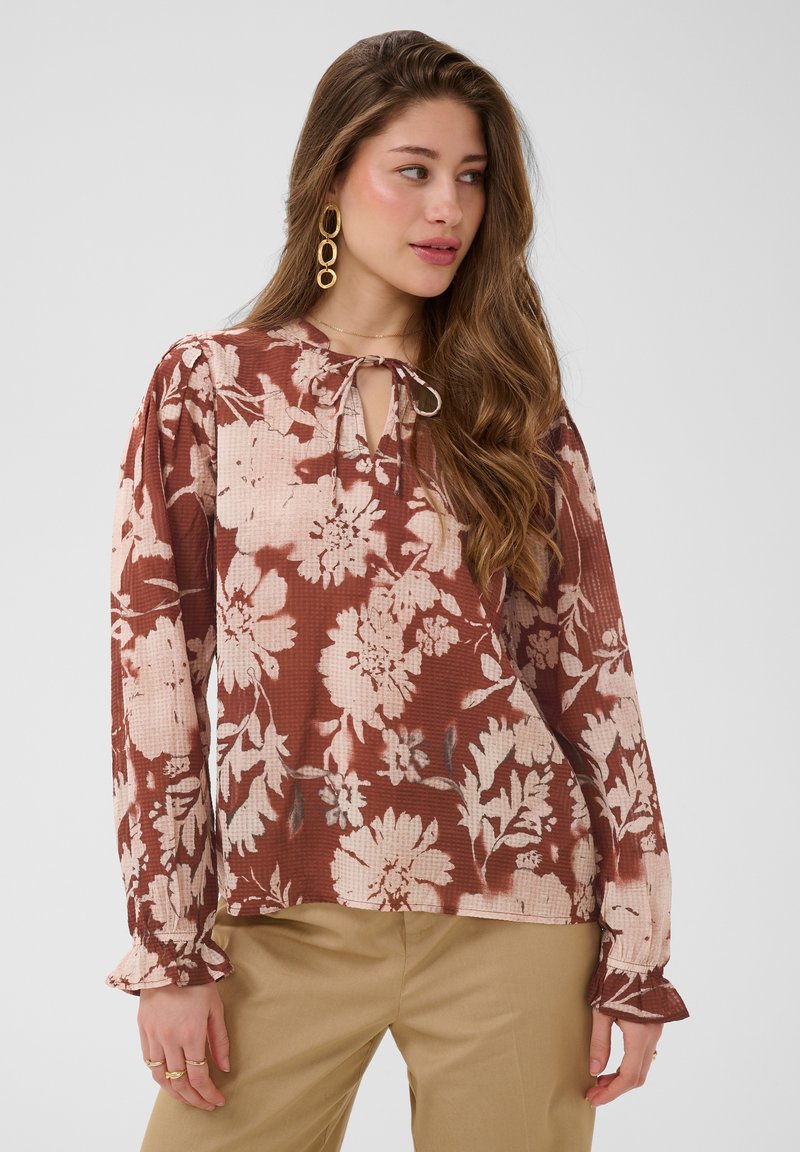 Femme aux longs cheveux bruns portant des boucles d’oreilles créoles en or et un chemisier marron à manches longues à motif floral associé à un pantalon beige, regardant sur le côté.