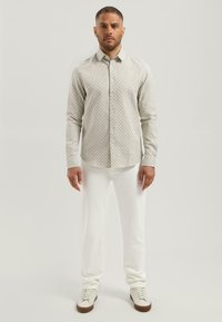 Man staat op een effen achtergrond en draagt een beige, gemêleerd, langemouwen overhemd met knopen, witte broek en witte sneakers met bruine zolen.