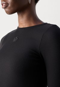 Primo piano di una persona che indossa una maglietta nera a coste con un piccolo logo nero ricamato vicino all'apertura del collo e un orecchino a cerchio d'argento.