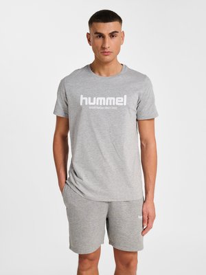 Unge mand står iført en grå hummel t-shirt og matchende grå shorts, hænderne afslappede, neutralt udtryk, ensartet hvid baggrund.
