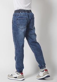 Denim jogger hlače u srednje plavoj boji, s elastičnim pojasom, manžetama na gležnjevima i dva stražnja džepa. Kombinirane s bijelim i crvenim tenisicama.