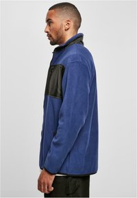 Giacca in pile blu con una tasca frontale nera e lucida. Presenta un colletto alto e chiusura con zip. Tessuto morbido, vestibilità regolare.