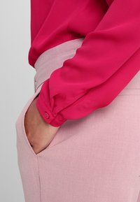 Blouse fuchsia à manches longues, avec des poignets à boutons, associée à un pantalon rose clair ajusté avec des poches latérales.