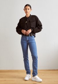 Černá zkrácená bomber bunda se dvěma předními kapsami, spojená se světlými modrými džínami a bílými teniskami na dřevěné podlaze proti neutrální zdi.