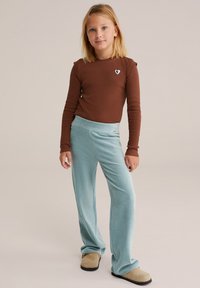 Brūna garām piedurknēm krekls ar mazu sirds detaļu, apvienots ar gaiši ziliem ribotiem platām kājām. Modelis valkā brūnas slip-on kurpes.