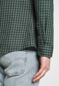 Camisa de cuadros verdes y azul marino con puños abotonados, material de algodón, dobladillo curvado y abertura lateral, combinada con jeans de mezclilla claros.