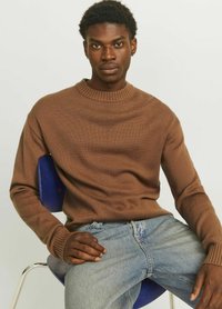 Brauner, gestrickter Pullover mit geripptem Rundhalsausschnitt und langen Ärmeln, kombiniert mit hellblauen Jeans, sitzt auf einem blauen Stuhl.