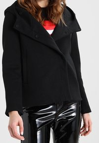 Sort dobbeltradet jakke med en stor hætte, som har en glat overflade og forlommer, parret med blanke sorte bukser.