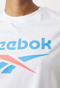 Bílý bavlněný tričko s velkým modrým logem "Reebok" a grafickým designovým prvkem v červené a modré barvě. Kulatý výstřih a hladká textura.