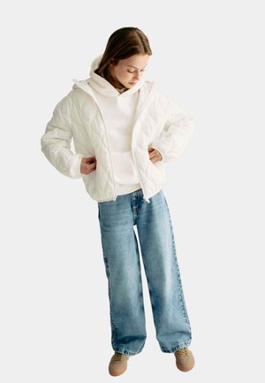 Vadderad vit jacka med huva, bärs över en vit sweatshirt, kombinerad med ljusblå vida jeans och bruna sneakers.