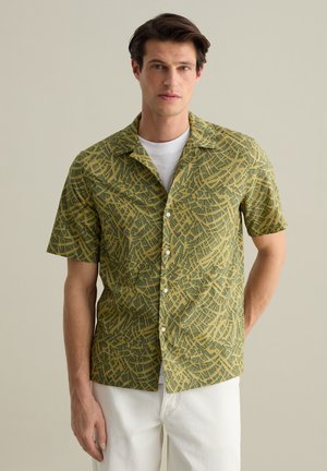 Camisa de manga corta en verde con un patrón geométrico amarillo. Presenta un cuello abierto, botones y un estilo de tejido ligero.
