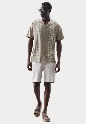 Man draagt een beige overhemd met korte mouwen en een verticale kettingprint, witte short, donkere zonnebril en beige sandalen met dubbele band loopt vooruit.