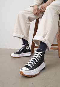 Person sitting on a wooden stool wearing beige rolled-up pants, black high-top sneakers with white laces, and dark socks on a grey floor.