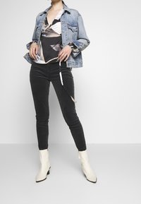 Denim jasje met een lichtblauwe wassing, zwarte slim-fit broek en witte puntige enkellaarsjes. Het shirt heeft een zwart en crème abstract patroon.