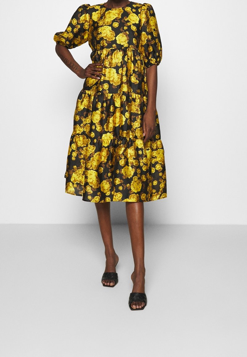 Femme portant une robe noire longueur genou avec un motif floral jaune et des manches bouffantes mi-longues, associée à des sandales noires à talons hauts.