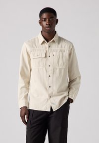 Off-white long-sleeve shirt in a lightweight fabric with two front pockets, button-up closure, and a small logo accent, paired with dark pants.