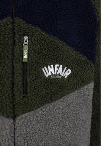 Veste polaire à blocs de couleurs bleu marine, olive et gris, fermeture éclair noire et logo "UNFAIR ATHLETICS" brodé sur la zone olive.