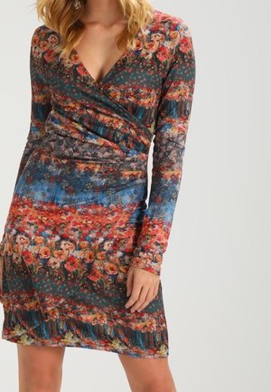 Vrouw die een jurk draagt met lange mouwen, een V-hals en een lengte tot de knieën, met een kleurrijk bloemenpatroon in rood, blauw en groene tinten.