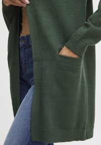Cardigan lavorato a maglia verde scuro con polsini a coste e tasche laterali, indossato sopra un top beige e abbinato a jeans blu. Texture morbida e design lungo.