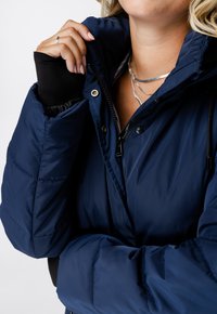 Marineblauwe pufferjas met een hoge kraag, ritssluiting aan de voorkant en elastische manchetten. Heeft een gladde textuur en gestikt patroon op de mouwen.