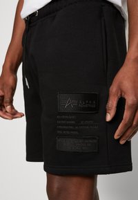 Czarne szorty Alpha Industries z naszywkami, noszone przez osobę, pokazujące szczegóły zamka błyskawicznego i sznurków na pasie.