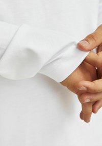 Balts garās piedurknes krekls ar gludu, teksturētu audumu. Piedurkne ir salocīta, parādot kārtīgas šuves un ciešu piegulšanu ap plaukstu.