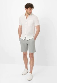 Camicetta bianca a maniche corte con taschino sul petto, abbinata a pantaloni corti grigi. Modello che indossa sneaker bianche, in piedi con le mani nelle tasche.