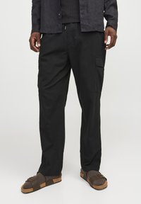 Pantalones cargo negros hechos de una tela ligera, con bolsillos laterales y un ajuste relajado. Combinados con sandalias marrones y una camiseta oscura.