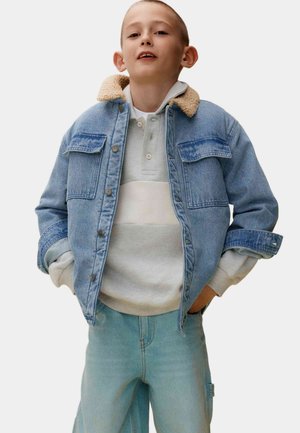 Veste en jean avec un col en shearling, deux poches poitrine, portée sur un polo rayé gris clair et blanc, associée à un jean bleu clair.