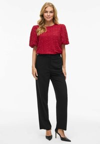Haut court à manches courtes texturé rouge associé à un pantalon noir ajusté et des talons hauts noirs. Le haut présente un motif ondulé pour plus de détails.