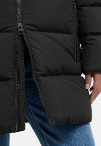 Giacca a vento nera con imbottitura spessa, dotata di chiusura a zip e texture morbida. Indossata sopra jeans in denim blu, che mostrano un fit casual.
