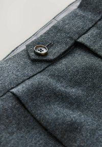 Grauer Stoff mit strukturierter Oberfläche, ausgestattet mit einem einzelnen, runden, schwarzen Knopf und einem dezenten Taschenklappen-Detail. Glatte Innenfutter.