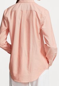 Světle růžová košile na knoflíky s dlouhými rukávy, límečkem a zakřiveným spodním lemem. Vyrobena z měkké, texturované látky se zadním skladem.