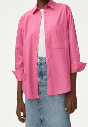 Persona que lleva una camisa rosa brillante abotonada sobre una camiseta blanca, combinada con una falda vaquera azul claro.