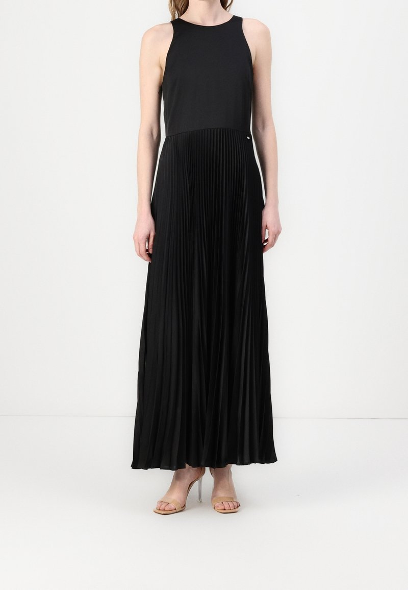 Femme portant une robe maxi plissée noire sans manches avec des sandales à talons hauts couleur nude sur un fond blanc.