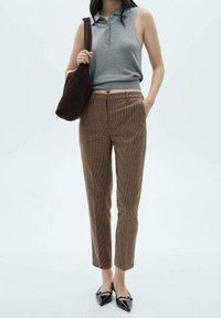 Un polo sans manches gris associé à un pantalon taillé à carreaux marron. Des chaussures noires à bouts pointus et un sac à épaule marron complètent le look.