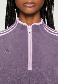 Lila fleece-pullover med hög krage, framdragkedja och tre vita ränder på axlarna. Har en broderad svart logo på bröstet.