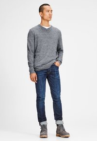Grauer V-Ausschnitt-Pullover, dunkelblaue Jeans, an den Säumen hochgekrempelt, und graue Stiefel. Der Pullover hat einen strukturierten Stoff; die Jeans sitzen eng an den Beinen.