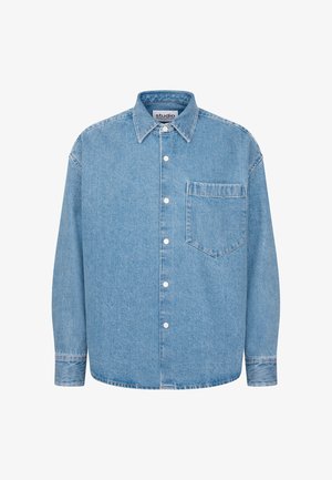 Jonge man in een lichtblauw oversized denim overhemd en spijkerbroek, staand tegen een effen lichte achtergrond met zijn handen in zijn zakken.