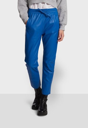 Person trägt leuchtend blaue Lederhose mit elastischem Bund und schwarzen Stiefeletten, steht vor einem schlichten Hintergrund.