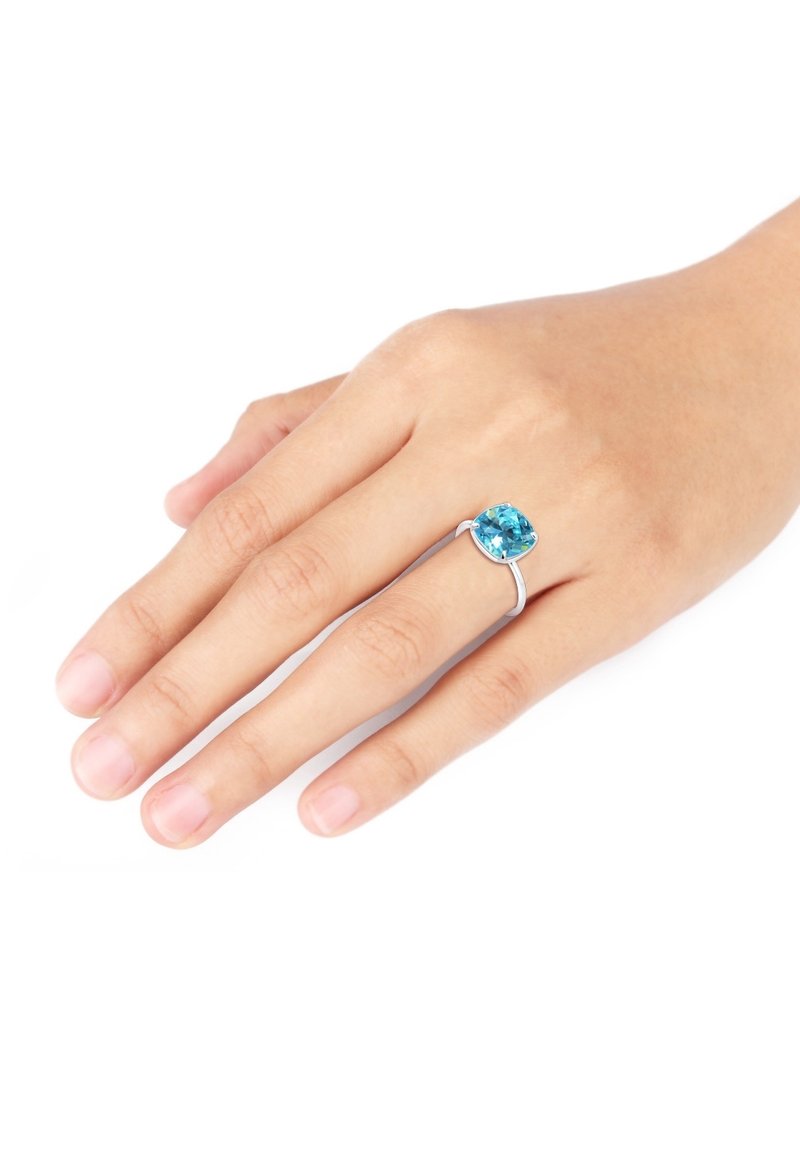 Main avec une bague en argent ornée d'une grande pierre précieuse carrée bleue portée à l'annulaire sur fond blanc.