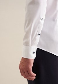 Nahaufnahme eines Arms einer Person, die ein weißes Langarmhemd mit schwarzen Knöpfen und einem kleinen schwarzen Rosenlogo nahe der Manschette trägt.
