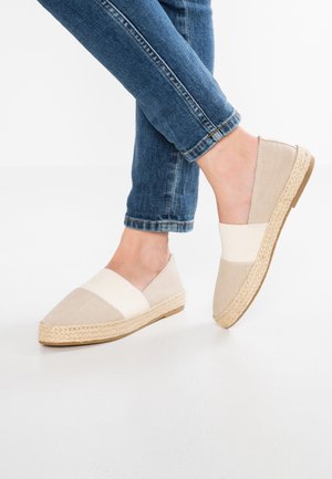Pieds portant des espadrilles beige et blanches sans lacets avec semelles en jute tressée, associées à un jean bleu retroussé sur fond blanc.