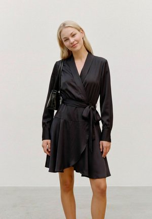 Une femme blonde présente une robe portefeuille noire nouée à la taille et un petit sac porté à l’épaule, sur un fond clair et uni.